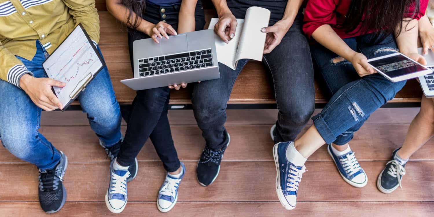 Students sitting on a bench using laptops and tablets for college financial aid research.