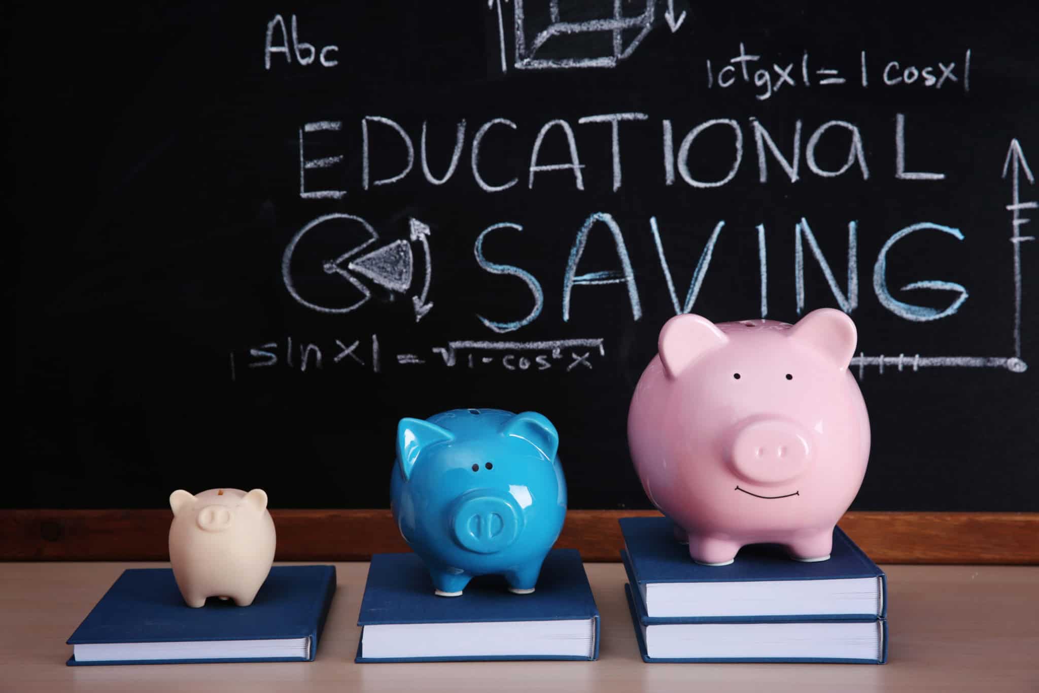 Three piggy banks of different sizes and colors on books in front of a chalkboard with educational '.